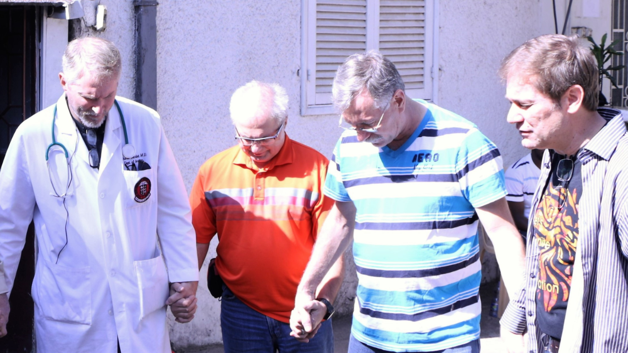 Four mission team members holding hands and praying together outdoors.