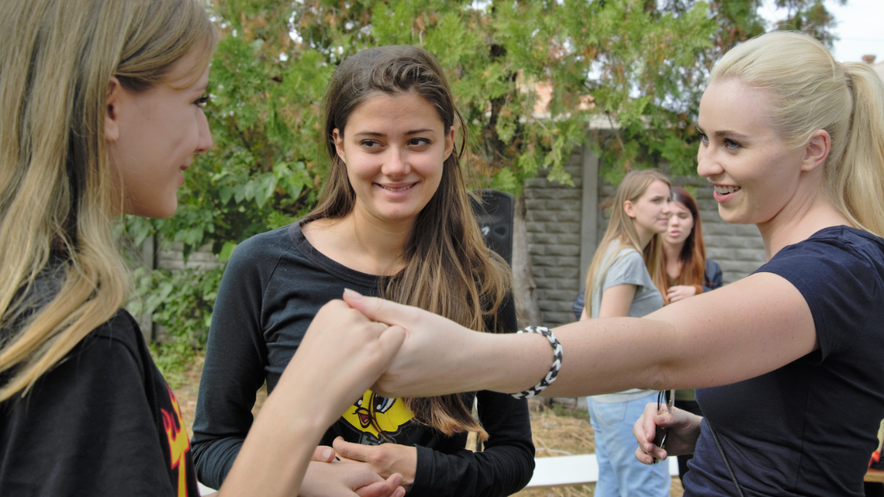 Three young women sharing a friendly fist bump during an outdoor gathering.